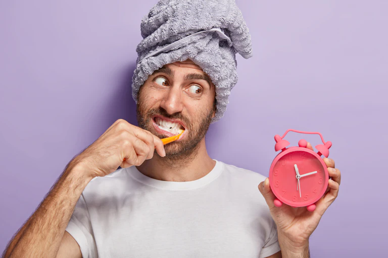 Man with towel on his head brushes his teeth while looking at a clock.