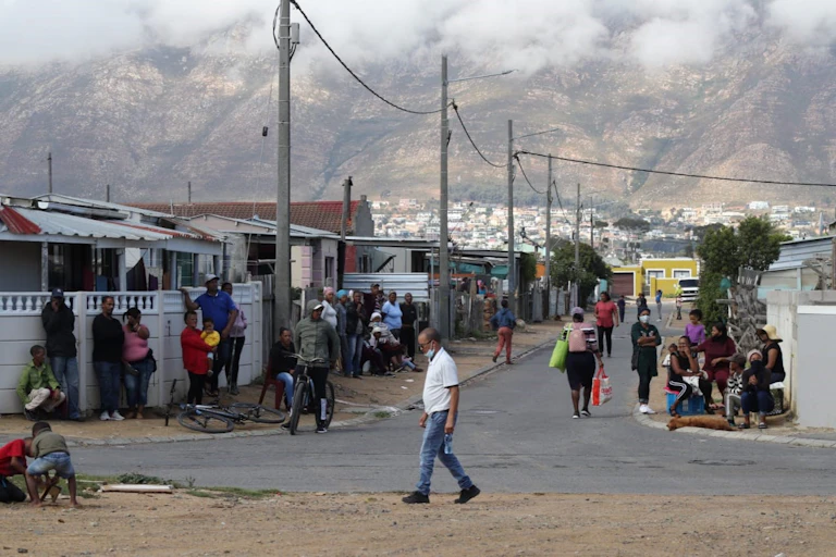 People stand outside yards in a street, with some hanging against a walled perimeter fence