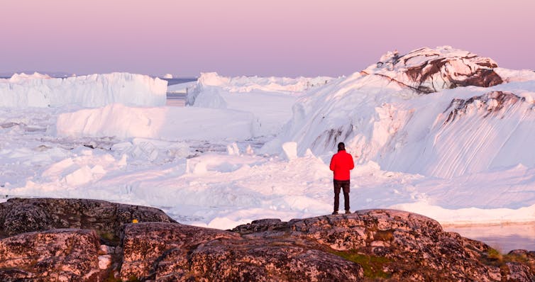 A scientist in red jacket stands before a glacier in Greenland.