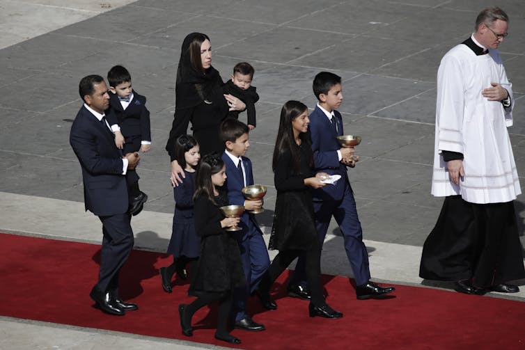 A woman wearing a black shawl walks down a red carpet at the Vatican with children around her.