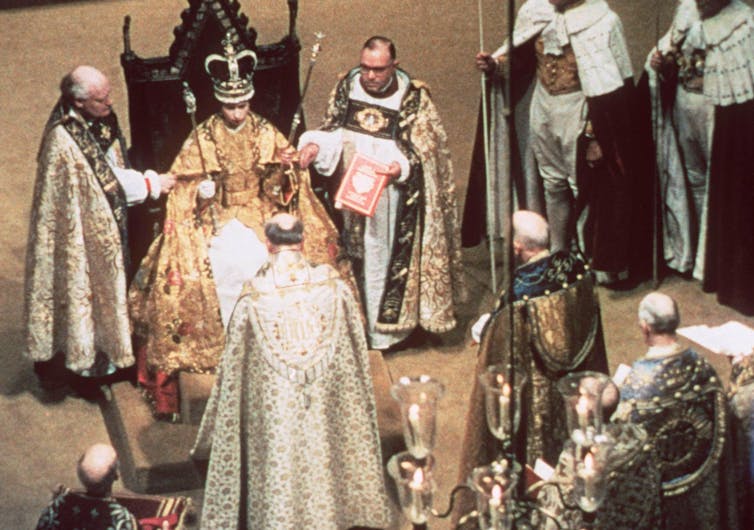 The Queen seated on a throne with full regalia, surrounded by bishops.