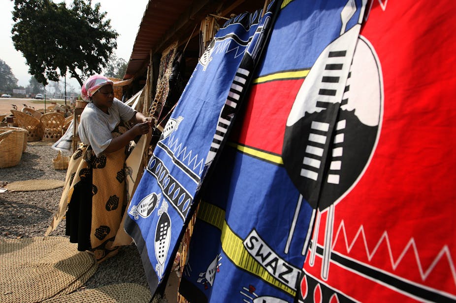 A woman outside a stall draped with cloths depicting shields and the word Swazi