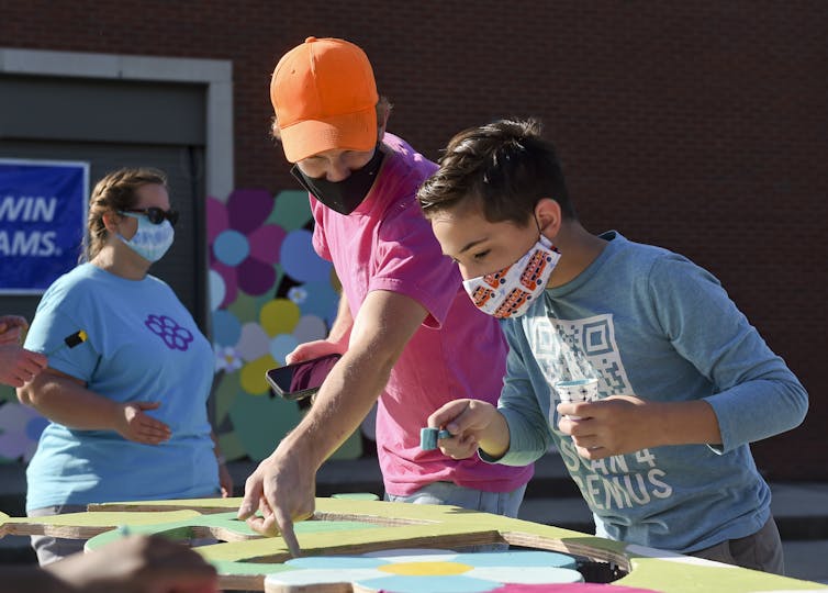 Boy wearing a blue shirt and face mask paints a piece of wood outside as a man wearing a pink shirt and orange baseball cap offers direction