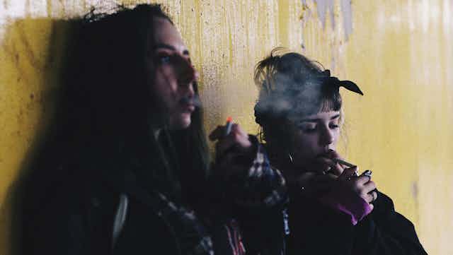 Deux adolescentes fument des cigarettes adossées à un mur.