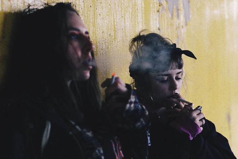 Deux adolescentes fument des cigarettes adossées à un mur.