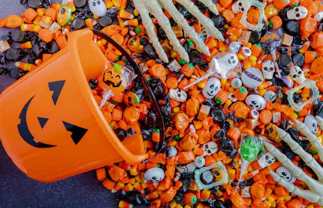 A jack o lantern bucket with candy spilling out and skeleton hands sorting it.