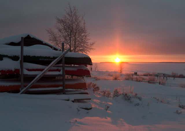 Canoes are stacked in the snow as the sun sets.