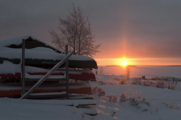 Canoes are stacked in the snow as the sun sets.