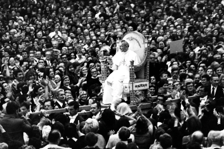 Pope John Paul I, dressed in white, rides on a platform through a crowd.