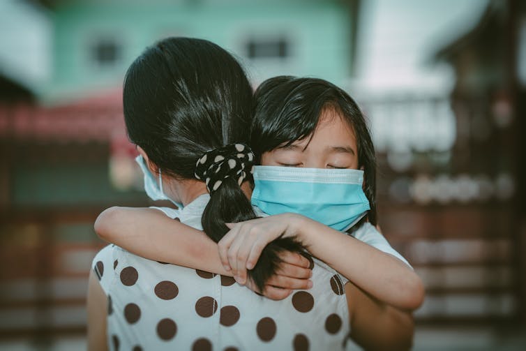 A young girl in a mask hugs her mother.