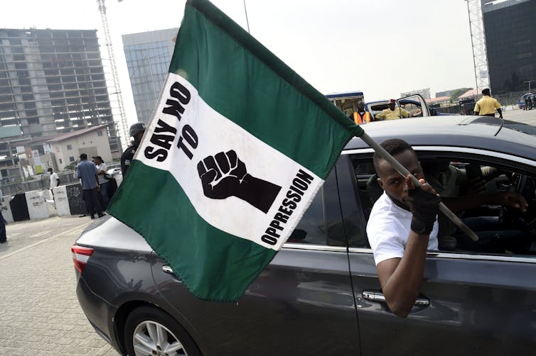 A man sticks his head out of a car, waving a large green-white-green flag.