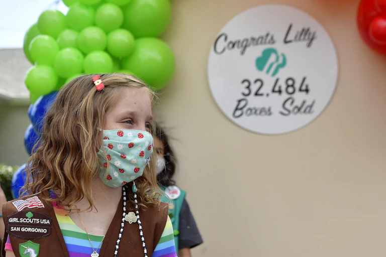 A Girl Scout wearing a mask and her uniform's vest celebrates her sale of 32,484 boxes of cookies.