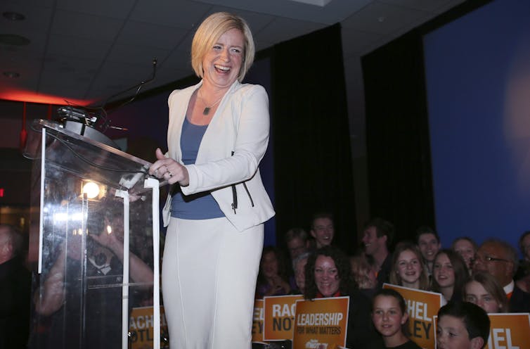 Rachel Notley, in a white suit and blue top, laughs from behind a podium.