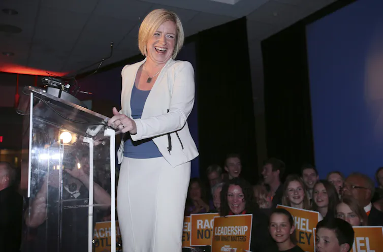 Rachel Notley, in a white suit and blue top, laughs from behind a podium.