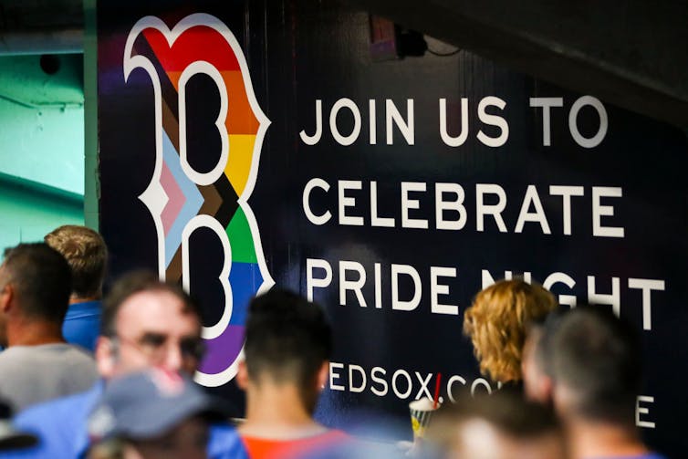 Fans walk past a Boston Red Sox logo with a Progress Pride flag superimposed over it.