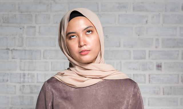 Woman wearing brown shirt and tan hijab stands against a grey brick wall
