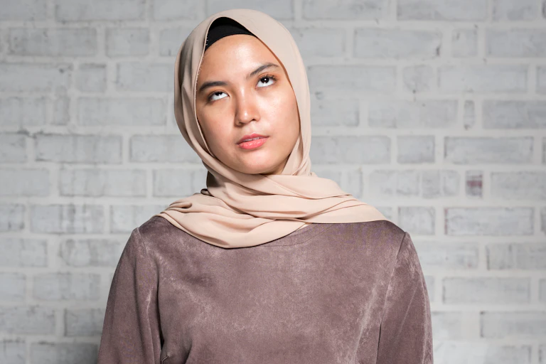 Woman wearing brown shirt and tan hijab stands against a grey brick wall