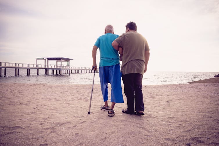 A younger man supports an older man walking on the beach.