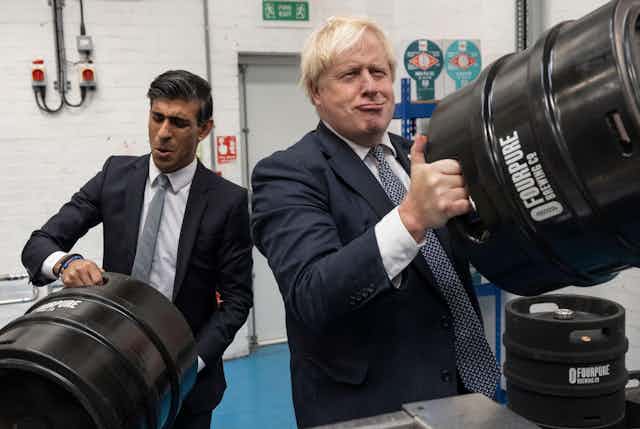 Rishi Sunak and Boris Johnson make faces as the pretend to lift heavy barrels of beer at a brewery.