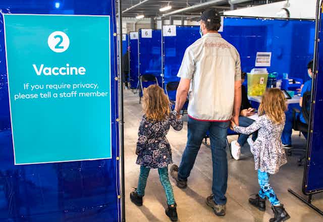 A man holding the hands of two girls is seen from behind, walking past a sign in a vaccine clinic