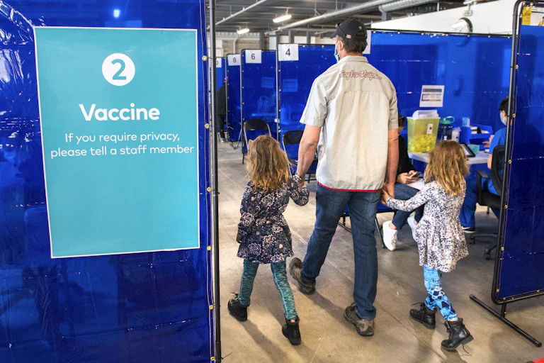 A man holding the hands of two girls is seen from behind, walking past a sign in a vaccine clinic