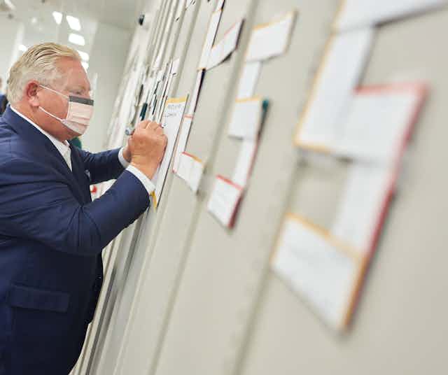 Doug Ford, wearing a mask, signs a thank you card to health-care workers pinned to a wall. 