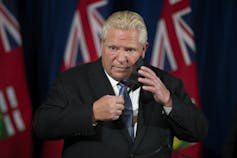A man in a suit and tie removes his mask as he starts a news briefing. Ontario flags are behind him.