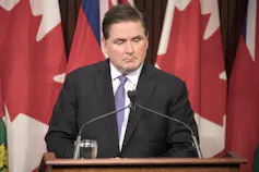 A man in a suit and tie stands behind a podium with Canadian flags behind him.