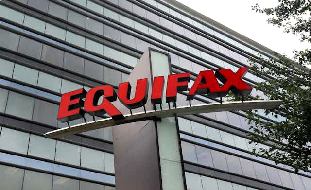 The glass façade of a large office building with a large sign mounted on a structure in front of the building bearing the name Equifax