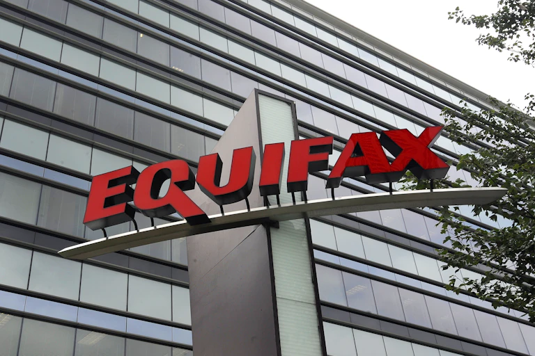 The glass façade of a large office building with a large sign mounted on a structure in front of the building bearing the name Equifax