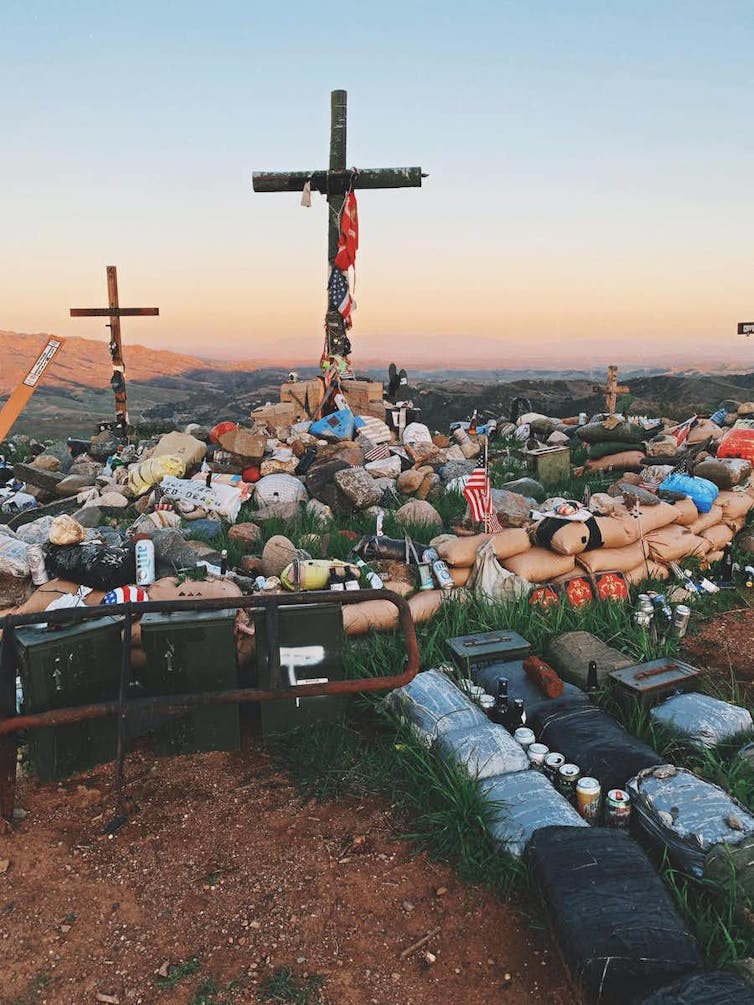 A cross stands on a hillside, with personal items like helmets, liquor bottles and insignia piled in the foreground.