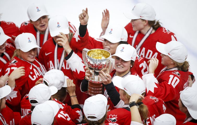 Women hockey players wearing white ball caps and red team Canada jersey's celebrate in a group as one woman holds up a trophy