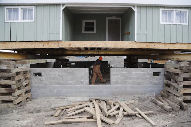 A house raised on piers with a new concrete block foundation being built beneath it.