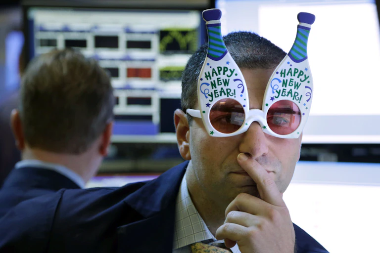 A man wearing white glasses in the shape of two champagne bottles protruding upwards from his eyes and with pinkish coloring over the lenses holds a finger to his lips