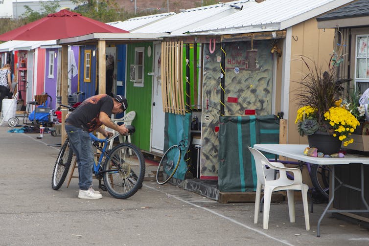 A man looks at his bike in front of a row of multi-coloured wooden cabins set up in a parking lot