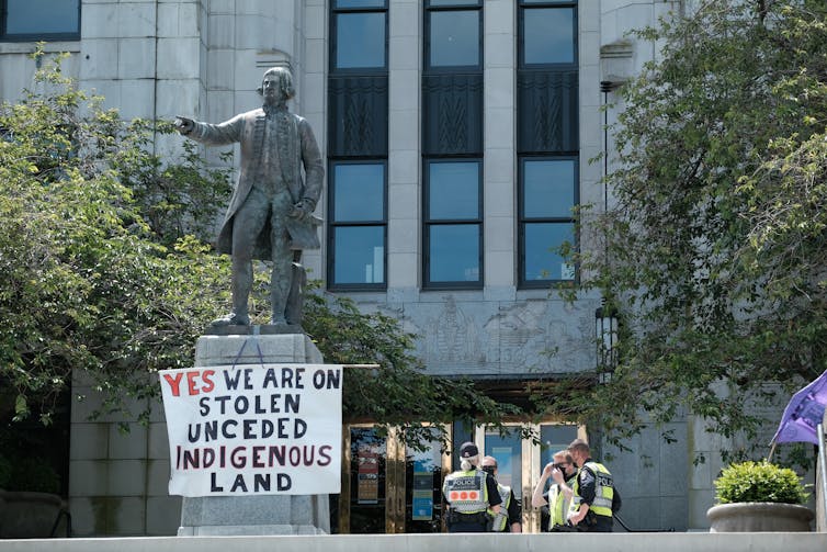 Vancouver Police officers talk behind a statue of George Vancouver covered in a sign recognizing Indigenous land rights