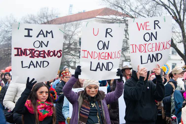 People hold signs reading 'I am on Indigenous land' 'U R on Indigenous land' and 'We R on Indigenous land'