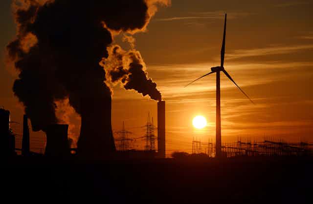 A wind turbine in front of smokestacks and power lines at sunset