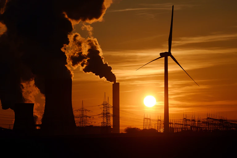 A wind turbine in front of smokestacks and power lines at sunset