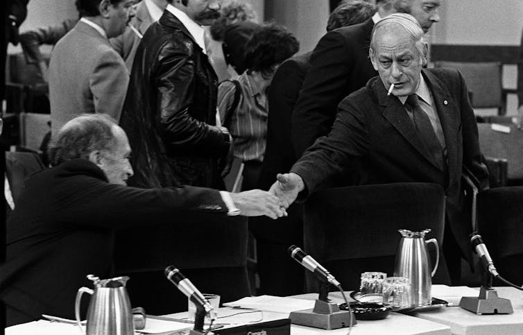 A man in a suit reaches out to shake hands with another man who has a cigarette dangling from his lips.