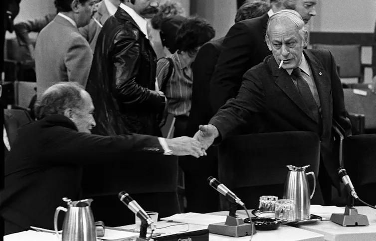 A man in a suit reaches out to shake hands with another man who has a cigarette dangling from his lips.
