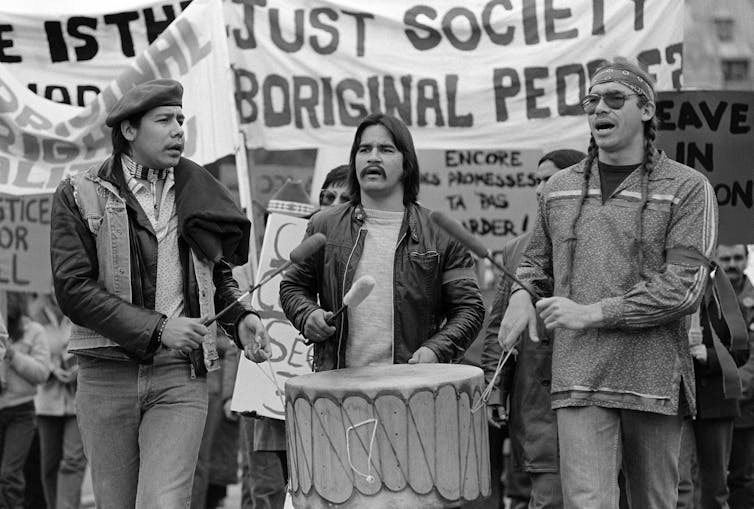 Three men with signs calling for a just society march in a line, banging on a drum.