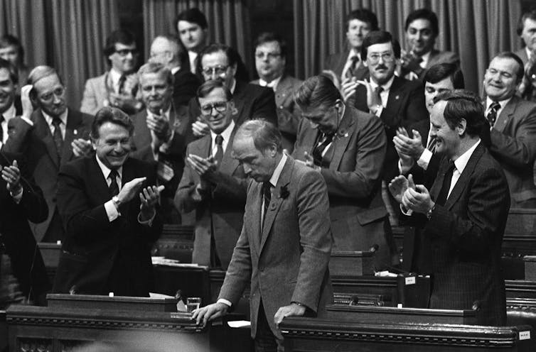 A man in a suit with a rose in his lapel bows his head while men around him applaud.