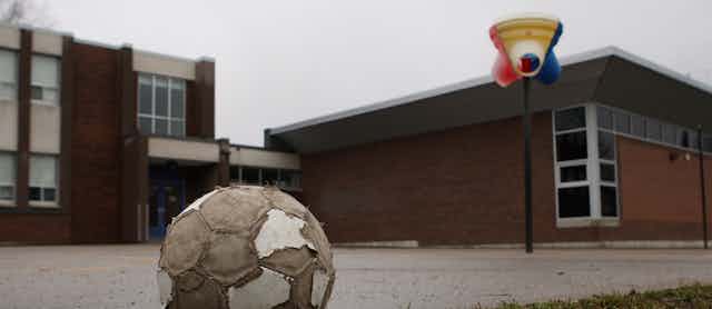 A tattered looking soccer ball is seen on pavement in a school yard with a lot of pavement.