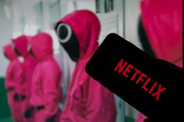 A row of Squid Game chharacters in pink tracksuits with black masks and a Netflix logo
