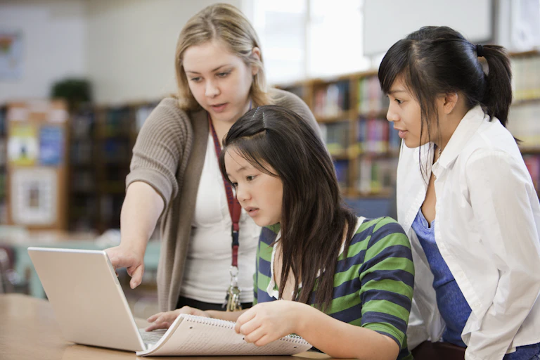 School librarian assists two students working on a laptop in library