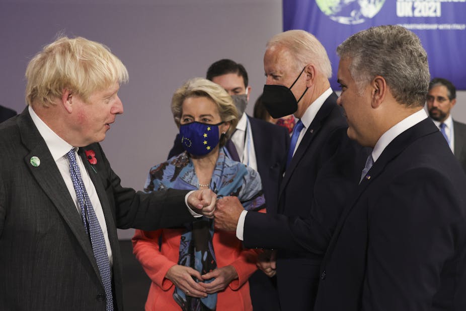 Boris Johnson and Joe Biden bumping fists at COP26 while Ursule von der Leyen looks on.