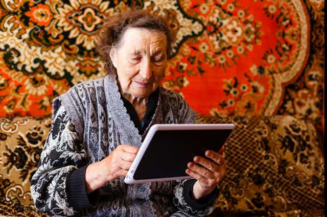 an older woman holds a tablet with a carpet in the background