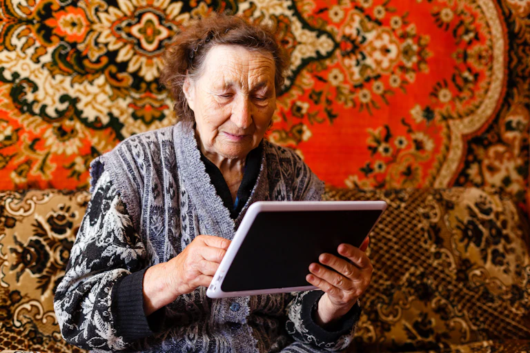 an older woman holds a tablet with a carpet in the background