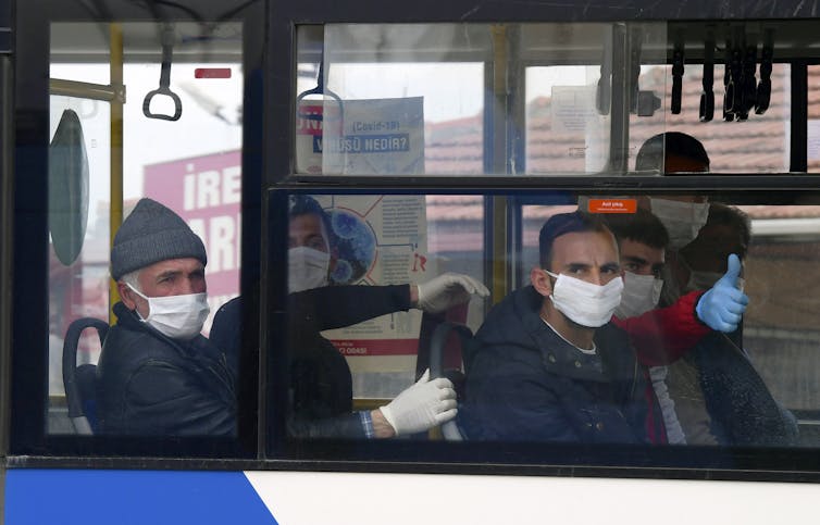 Men wearing masks and plastic gloves sit in a bus. One gives the thumb's up sign.